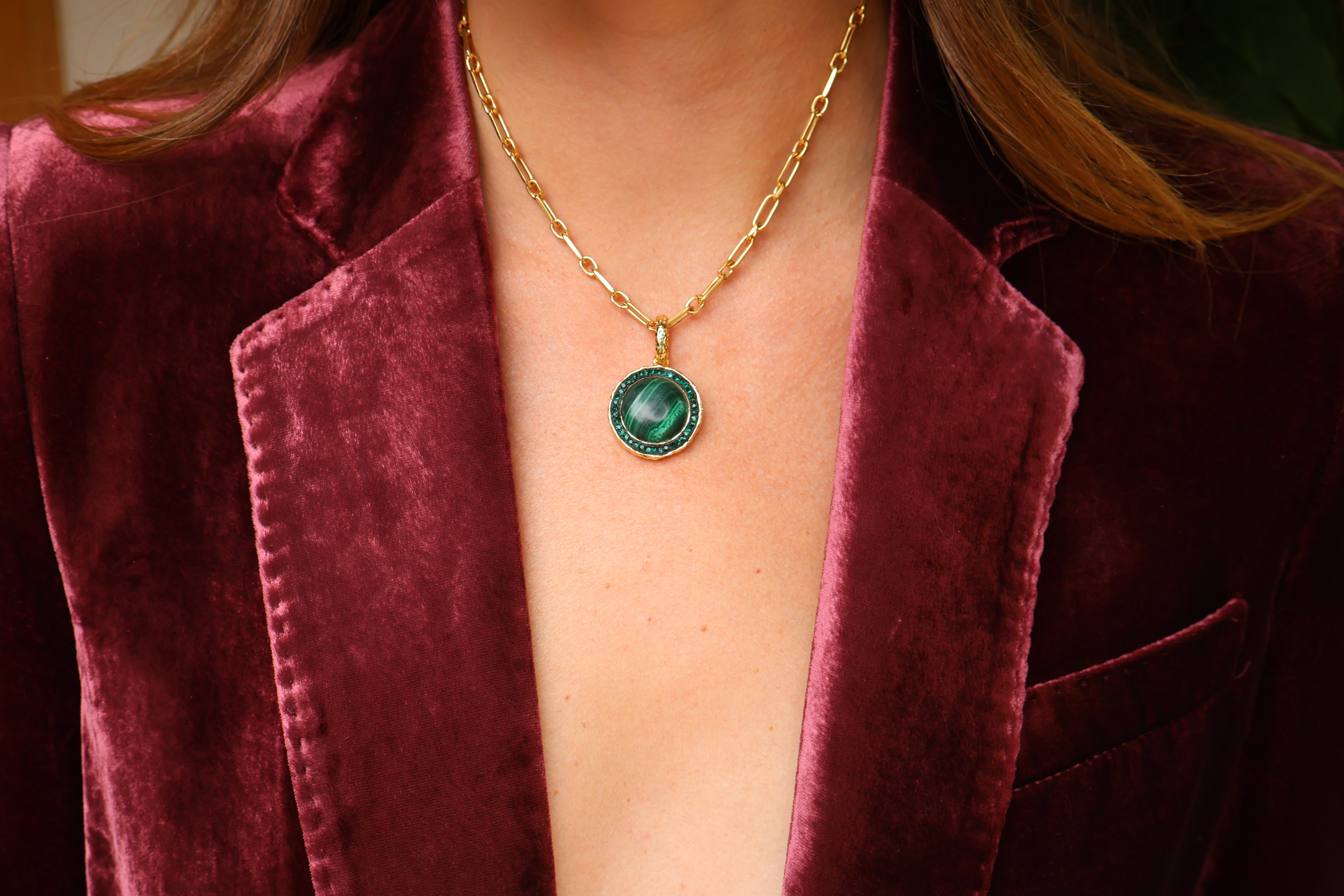A gold chain necklace featuring a smooth, round green malachite pendant surrounded by sparkling crystals, styled beautifully with a luxurious burgundy velvet blazer.