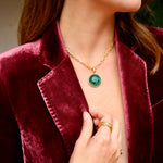 A close-up lifestyle shot of a woman wearing a gold chain necklace with a round green gemstone pendant and a green moon shaped earring, styled with a burgundy velvet blazer. 