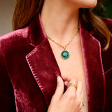 A close-up lifestyle shot of a woman wearing a gold chain necklace with a round green gemstone pendant and a green moon shaped earring, styled with a burgundy velvet blazer. 