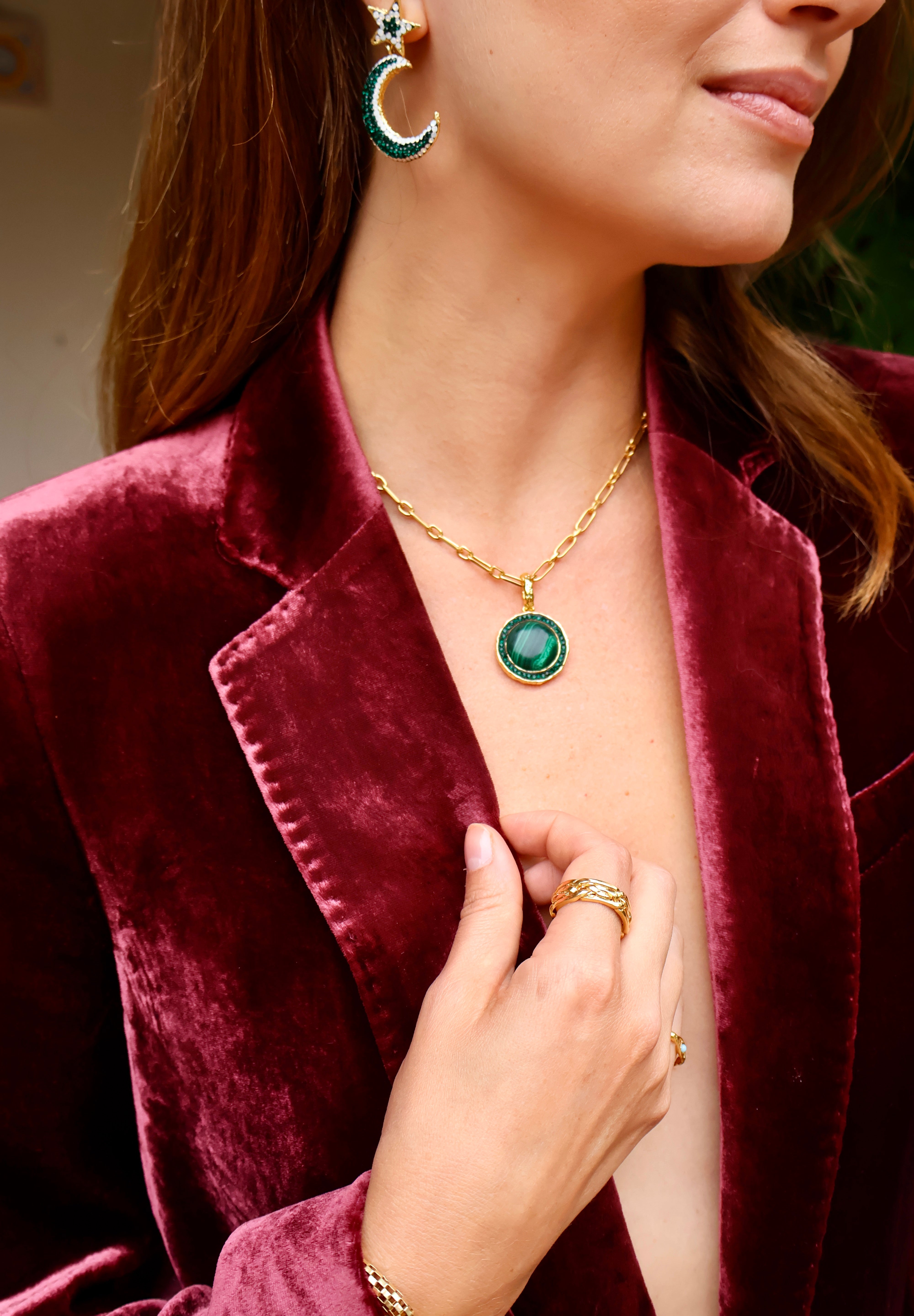 A close-up lifestyle shot of a woman wearing a gold chain necklace with a round green gemstone pendant and a green moon shaped earring, styled with a burgundy velvet blazer. 