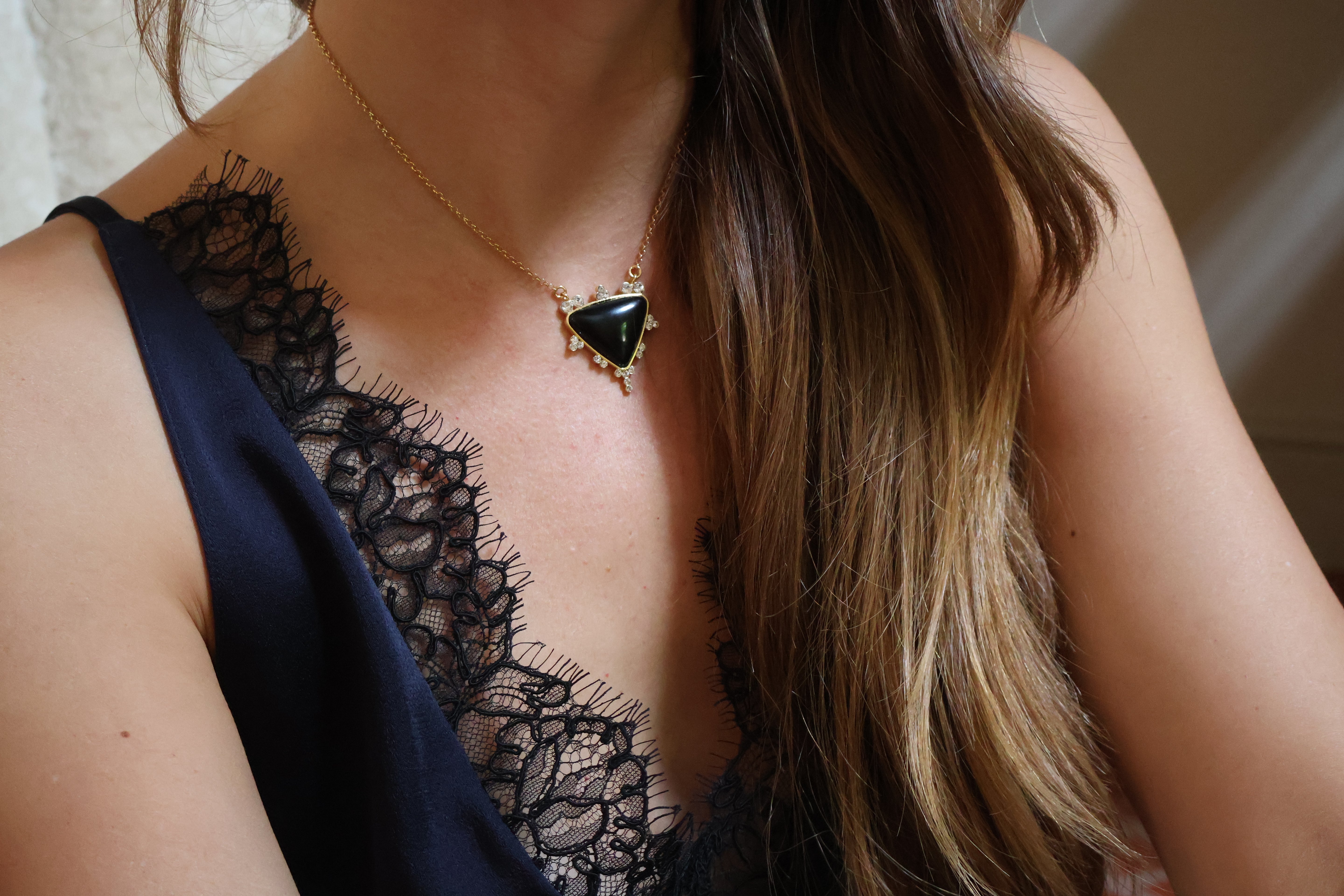 A close-up of a woman wearing a gold chain necklace with a triangular onyx pendant, styled with a black lace camisole top.