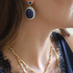 A close-up lifestyle shot of a woman wearing a gold and blue drop earring, layered gold necklaces, and a navy blue lace top.