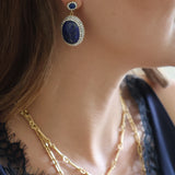 A close-up lifestyle shot of a woman wearing a gold and blue drop earring, layered gold necklaces, and a navy blue lace top.