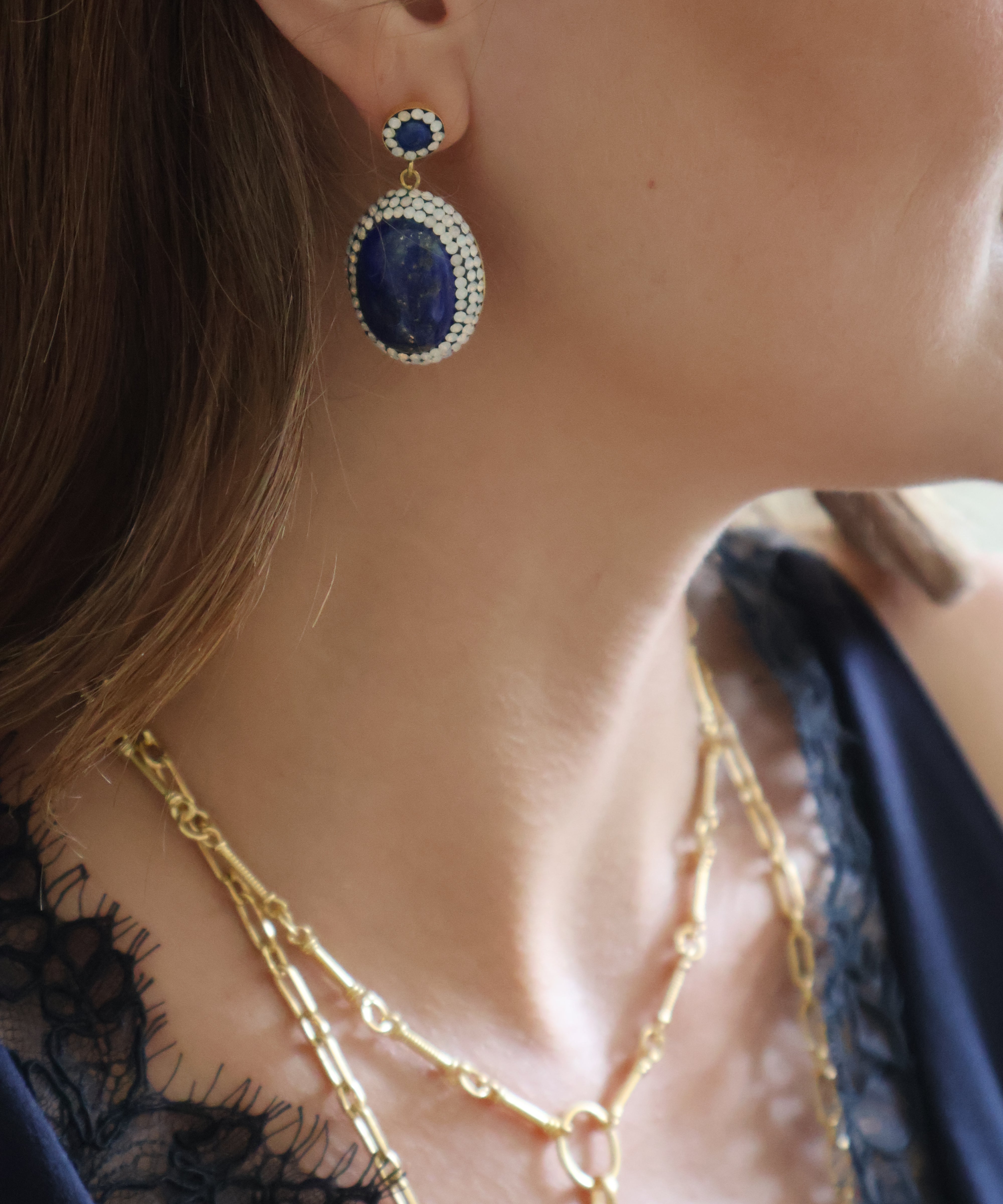A close-up lifestyle shot of a woman wearing a gold and blue drop earring, layered gold necklaces, and a navy blue lace top.