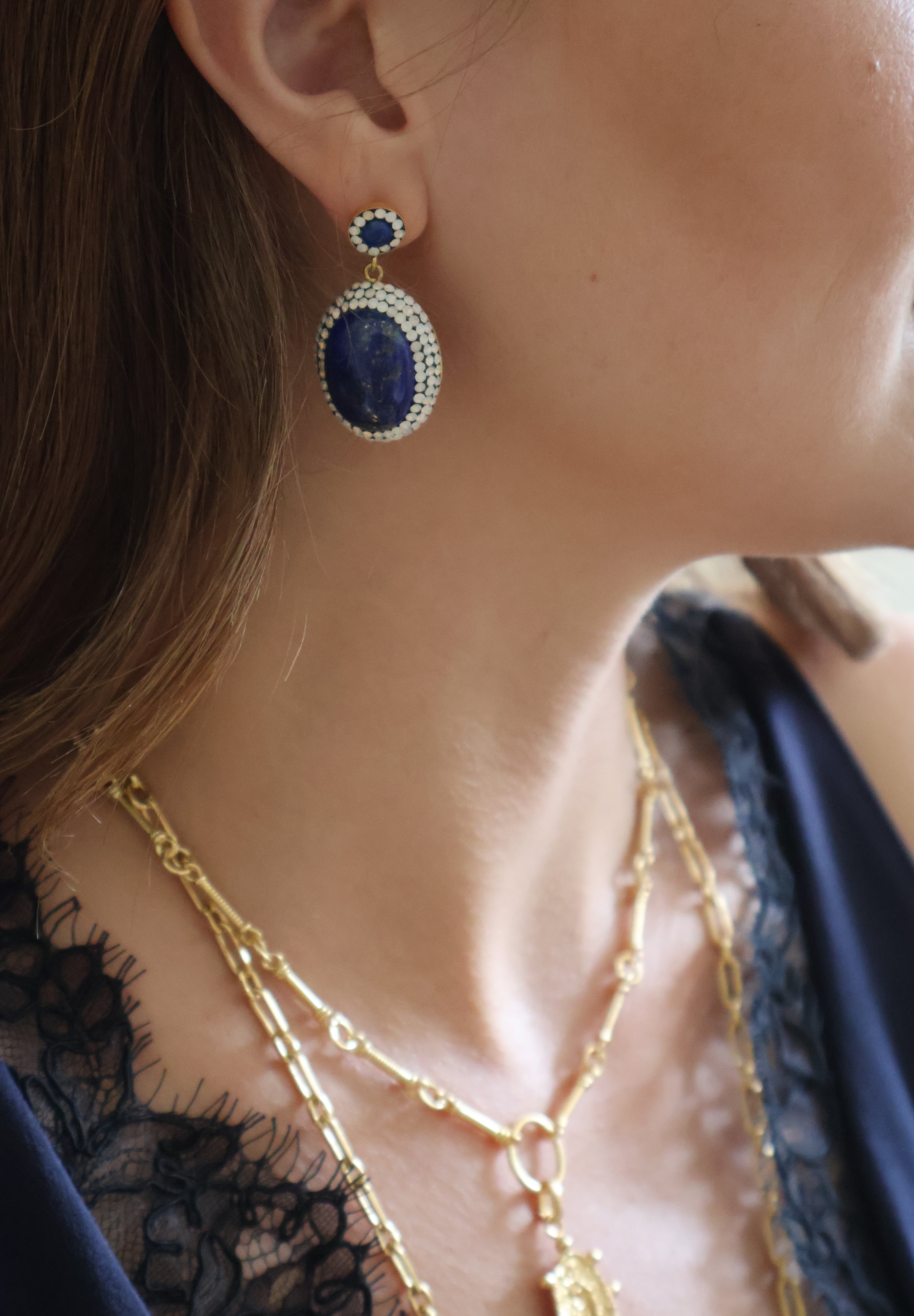 A close-up lifestyle shot of a woman wearing a gold and blue drop earring, layered gold necklaces, and a navy blue lace top.