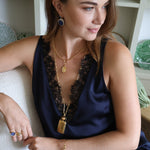 A woman wearing a navy blue satin slip dress with black lace trim, accessorized with gold necklaces, bracelets, and rings, sitting on a white textured chair in a bright room.