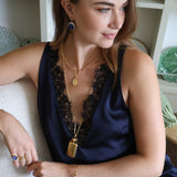A woman wearing a navy blue satin slip dress with black lace trim, accessorized with gold necklaces, bracelets, and rings, sitting on a white textured chair in a bright room.