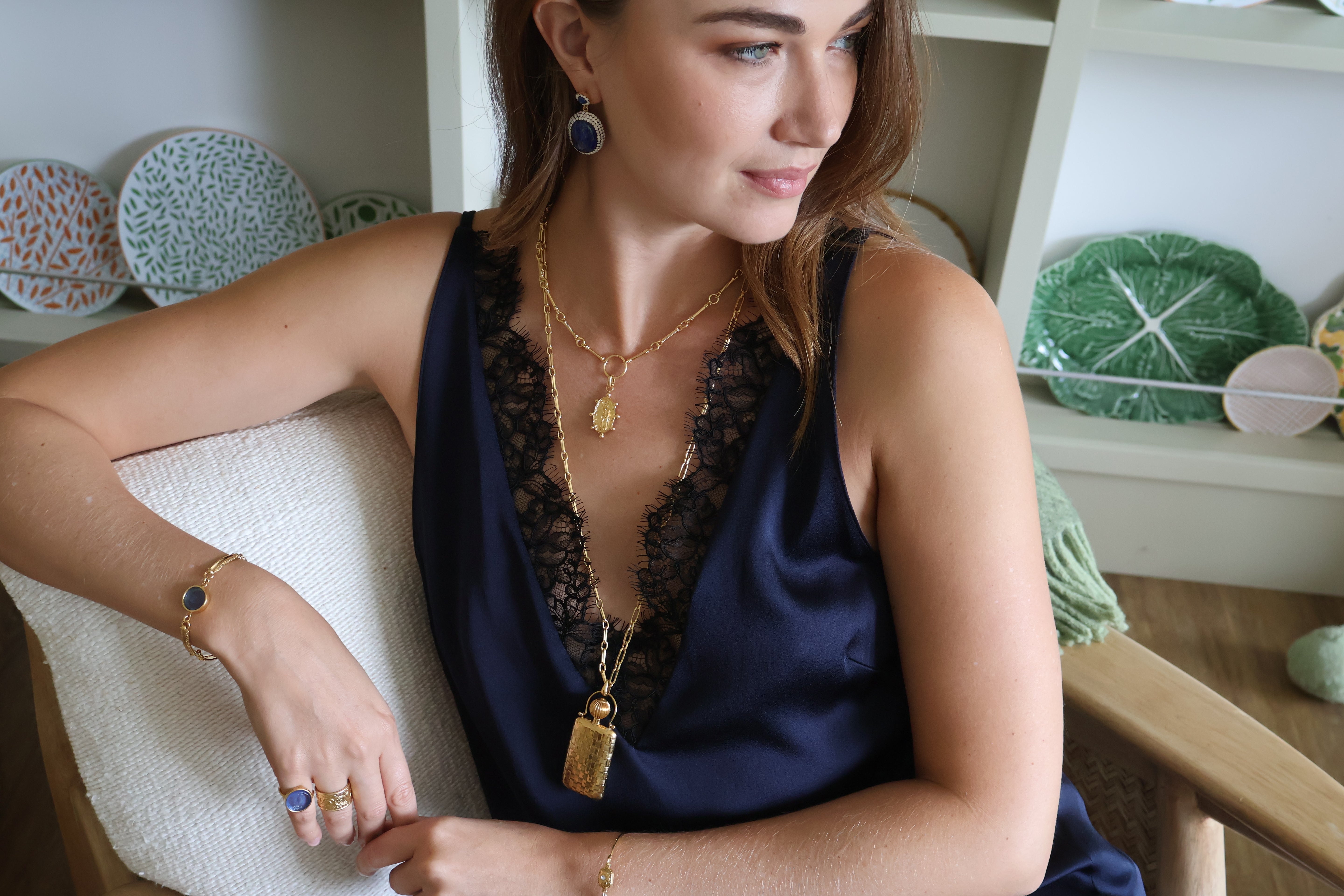 A woman wearing a navy blue satin slip dress with black lace trim, accessorized with gold necklaces, bracelets, and rings, sitting on a white textured chair in a bright room.