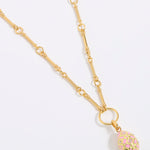 Gold necklace with a pink and gold egg shaped pendant on a light gray background