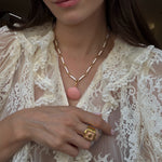 Close-up of a person wearing a delicate necklace with a pink pendant and gold ring, against a neutral background.