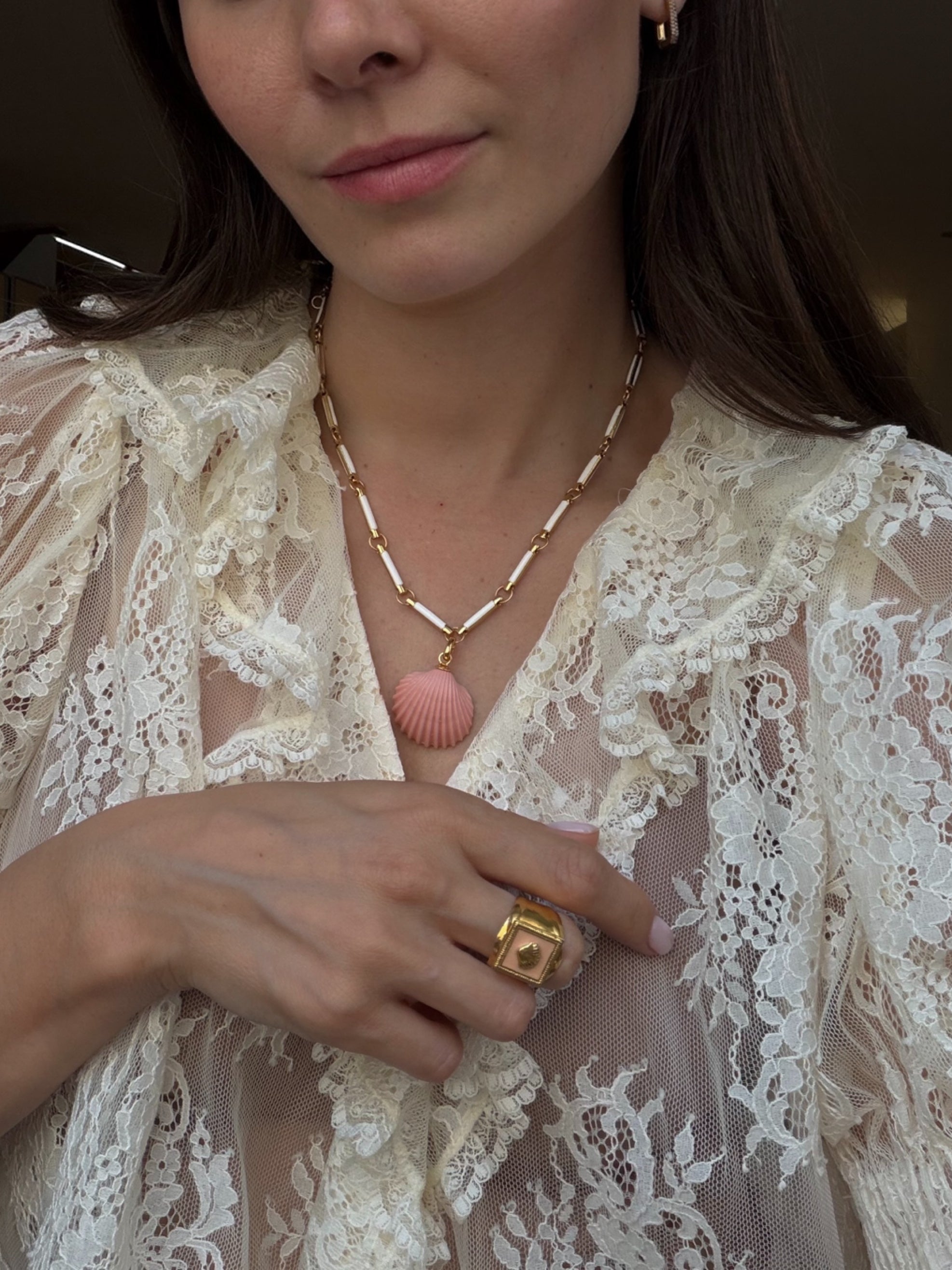 Close-up of a person wearing a delicate necklace with a pink pendant and gold ring, against a neutral background.