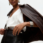 Woman wearing a brown leather jacket over a white top with silver cuff bracelet and statement silver earrings on a  light gray background