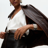 Woman wearing a brown leather jacket over a white top with silver cuff bracelet and statement silver earrings on a  light gray background