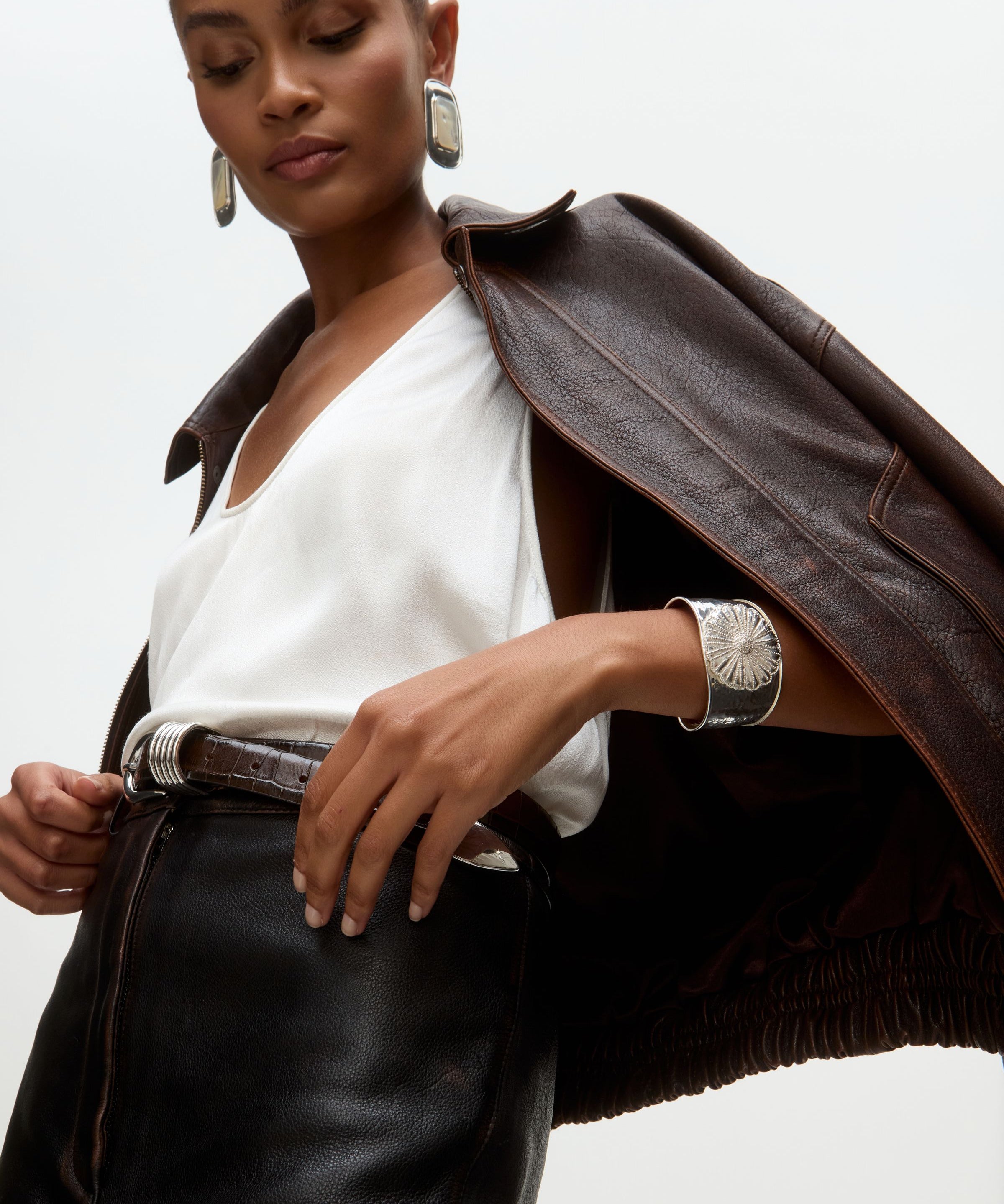 Woman wearing a brown leather jacket over a white top with silver cuff bracelet and statement silver earrings on a  light gray background