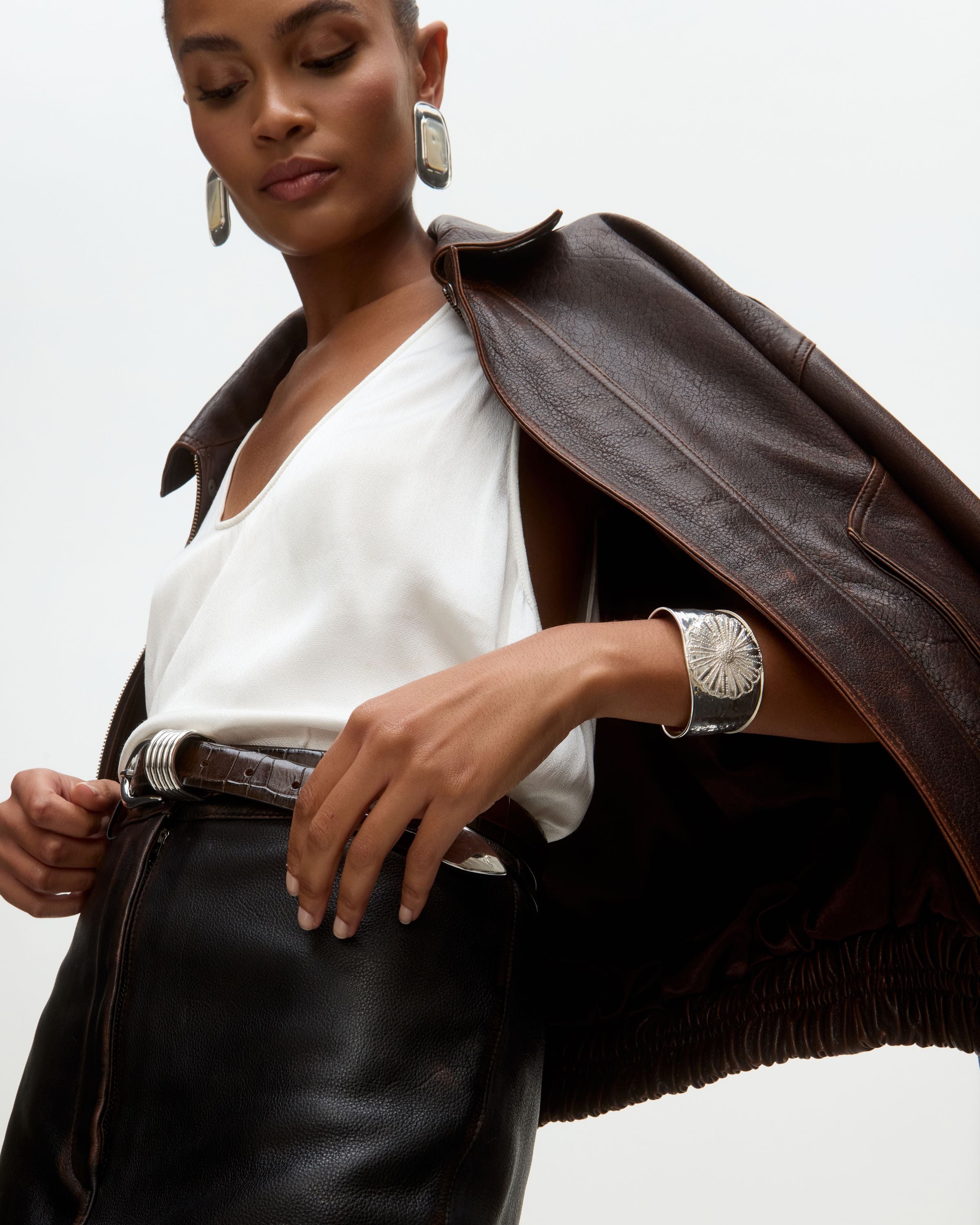 Woman wearing a brown leather jacket over a white top with silver cuff bracelet and statement silver earrings on a  light gray background