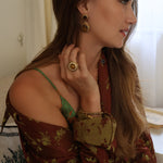 A woman in profile wearing a rust-brown floral off-the-shoulder top, tigers eye drop earrings, and a large gold ring with a tigers eye stone, in a softly lit indoor setting.