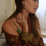 A woman in profile wearing a rust-brown floral off-the-shoulder top, tigers eye drop earrings, and a large gold ring with a tigers eye stone, in a softly lit indoor setting.