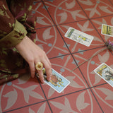 A person's hand resting on a red patterned tile floor next to several tarot cards, wearing a brown and green patterned robe and a gold ring.