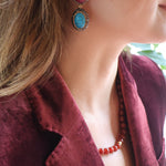 A close-up lifestyle shot of a woman wearing a pair of statement drop earrings. Each earring features a teardrop-shaped red ruby set in an ornate gold-toned setting, with an oval turquoise blue gemstone suspended beneath in a matching gold-toned setting.