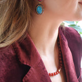 A close-up lifestyle shot of a woman wearing a pair of statement drop earrings. Each earring features a teardrop-shaped red ruby set in an ornate gold-toned setting, with an oval turquoise blue gemstone suspended beneath in a matching gold-toned setting.