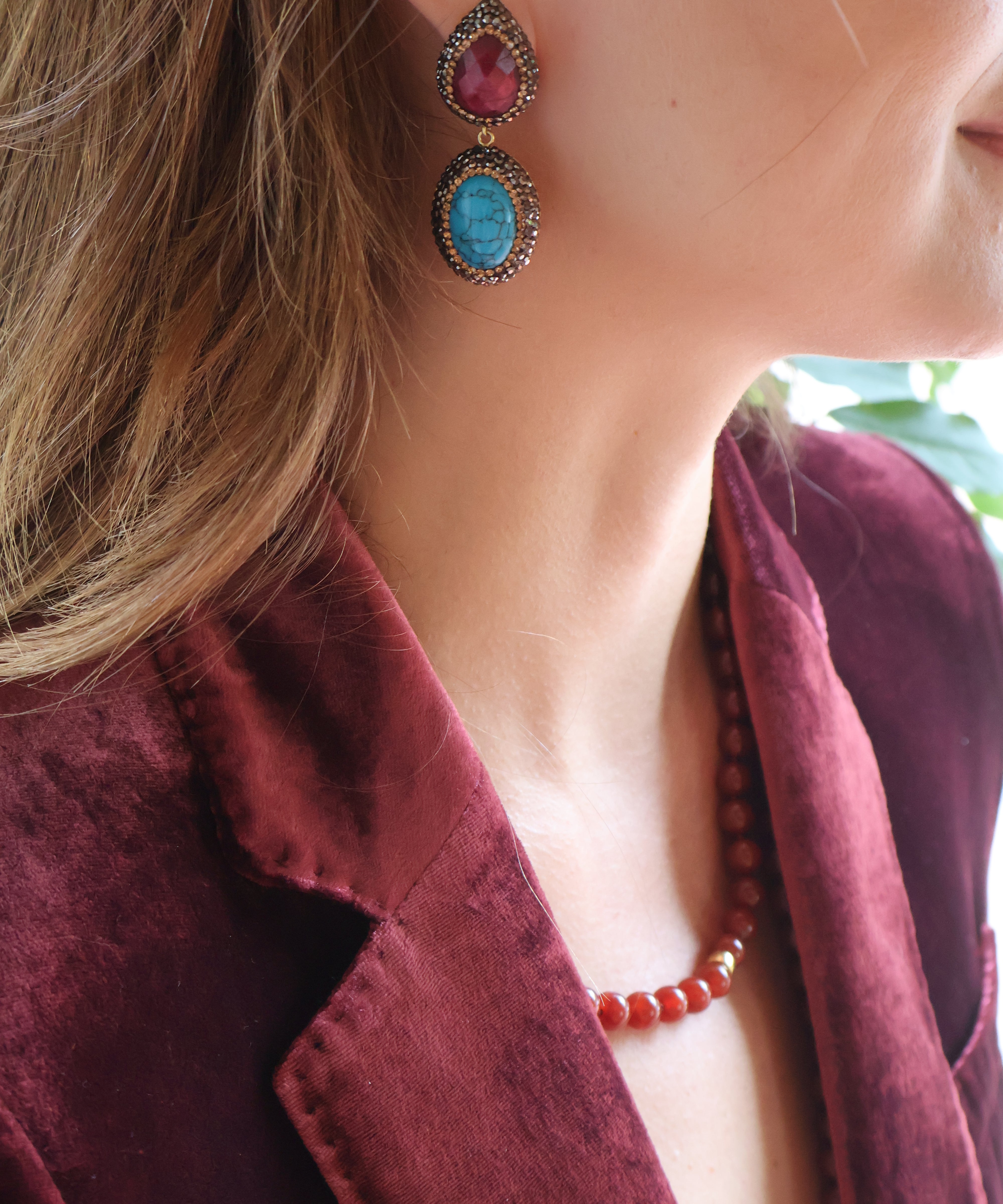 A close-up lifestyle shot of a woman wearing a pair of statement drop earrings. Each earring features a teardrop-shaped red ruby set in an ornate gold-toned setting, with an oval turquoise blue gemstone suspended beneath in a matching gold-toned setting.