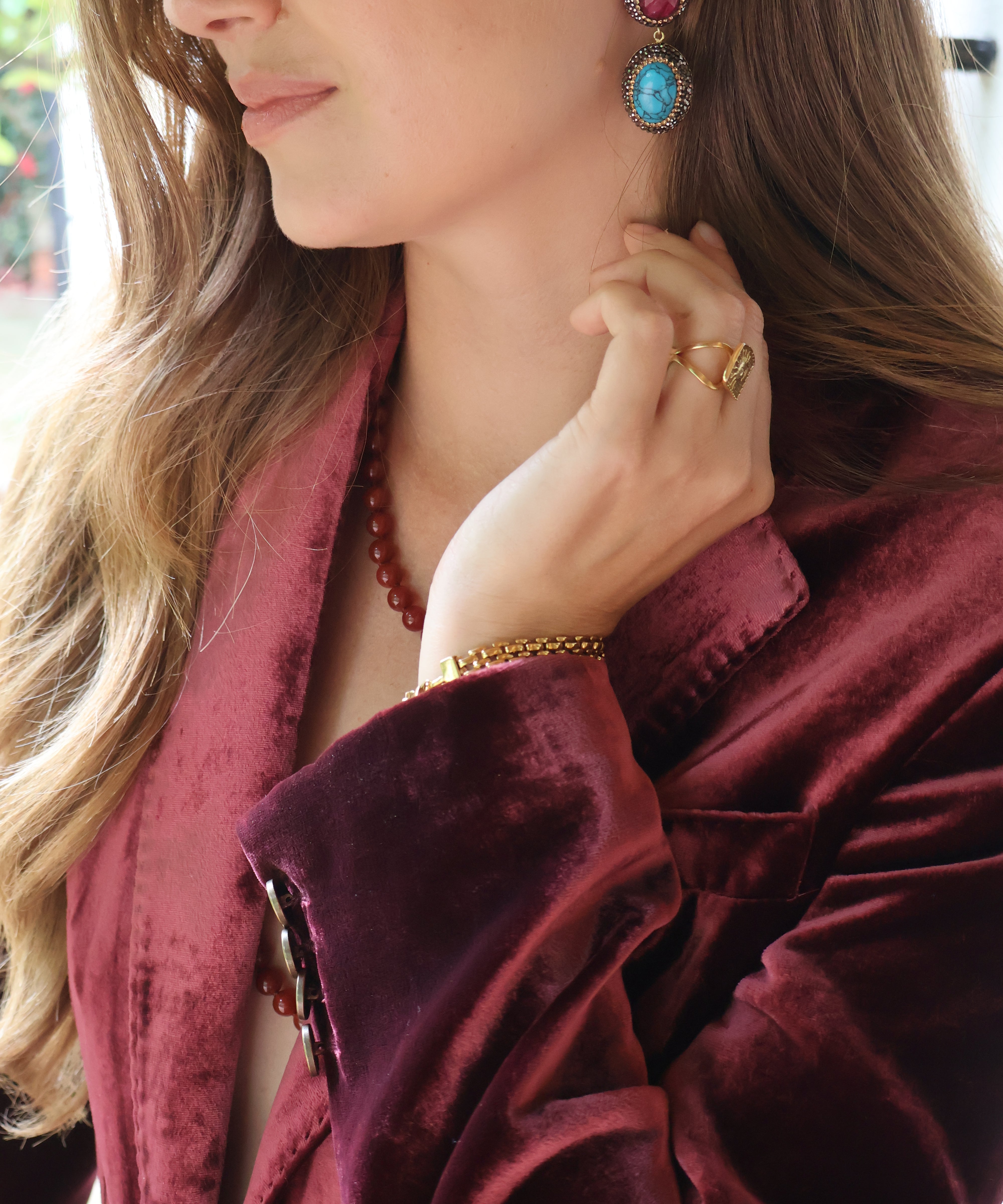 A close-up lifestyle shot of a woman wearing a deep burgundy velvet blazer, gold jewellery including a ring and bracelet, and a pair of statement earrings with red ruby and turquoise stones.