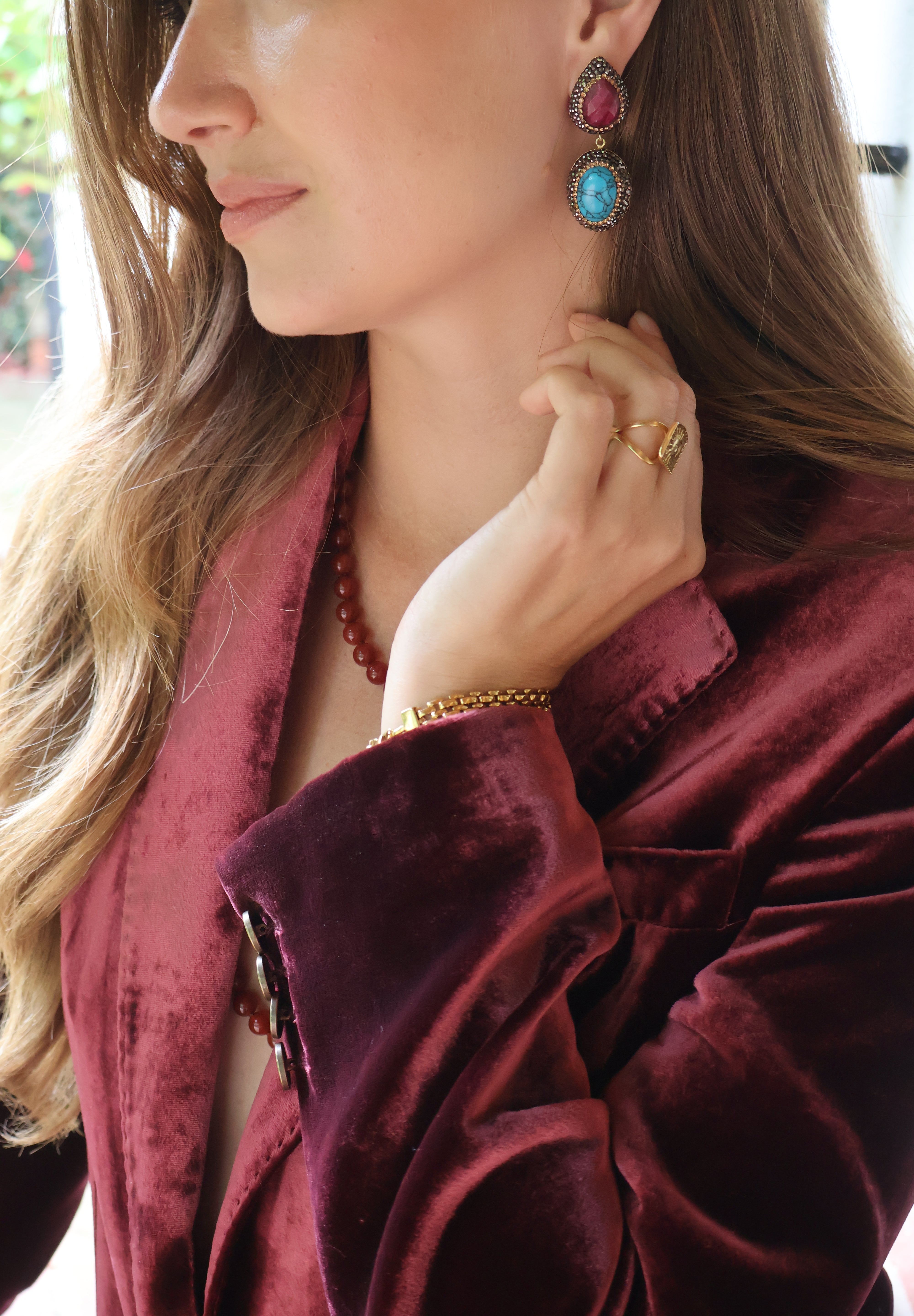 A close-up lifestyle shot of a woman wearing a deep burgundy velvet blazer, gold jewellery including a ring and bracelet, and a pair of statement earrings with red ruby and turquoise stones.