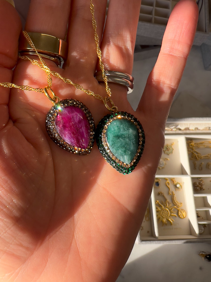 ruby and emerald pendant necklaces being held in a hand with natural sunlight shining on them both creating sparkle