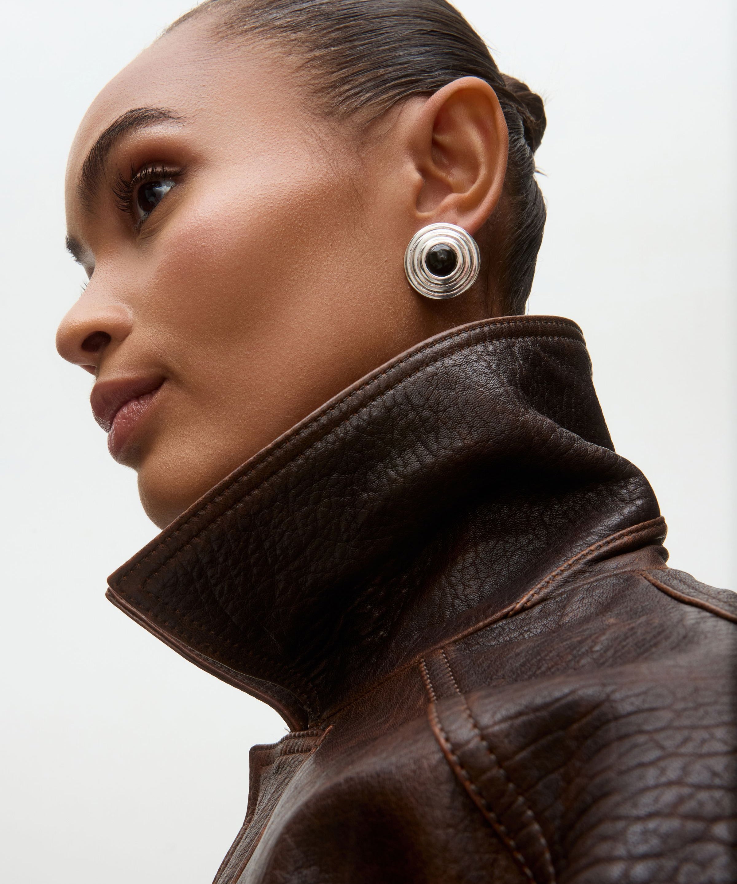 Woman wearing a brown leather jacket and silver earrings on a light gray background