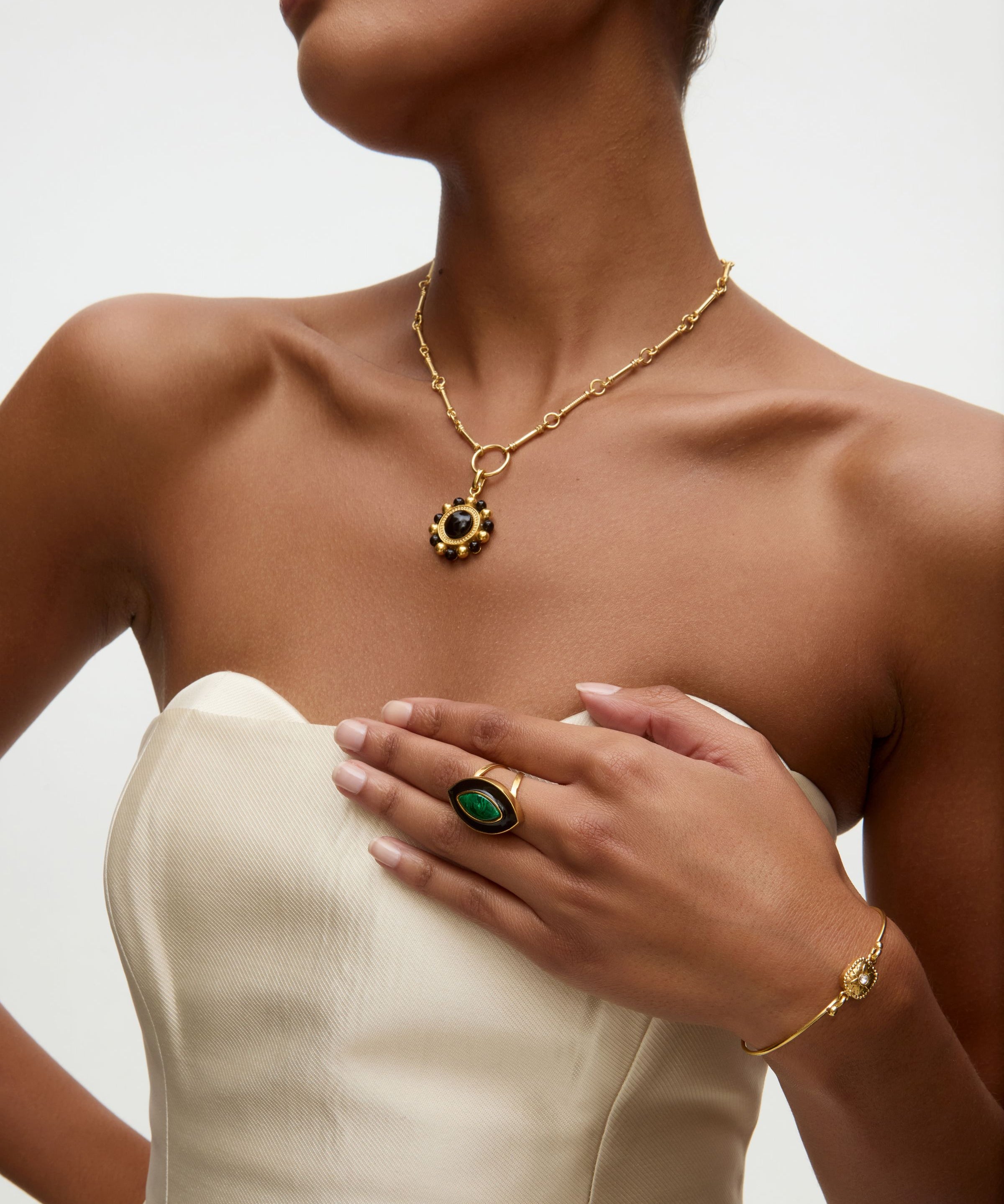 Woman wearing gold necklace and charm , green ring, and bracelet on a neutral background