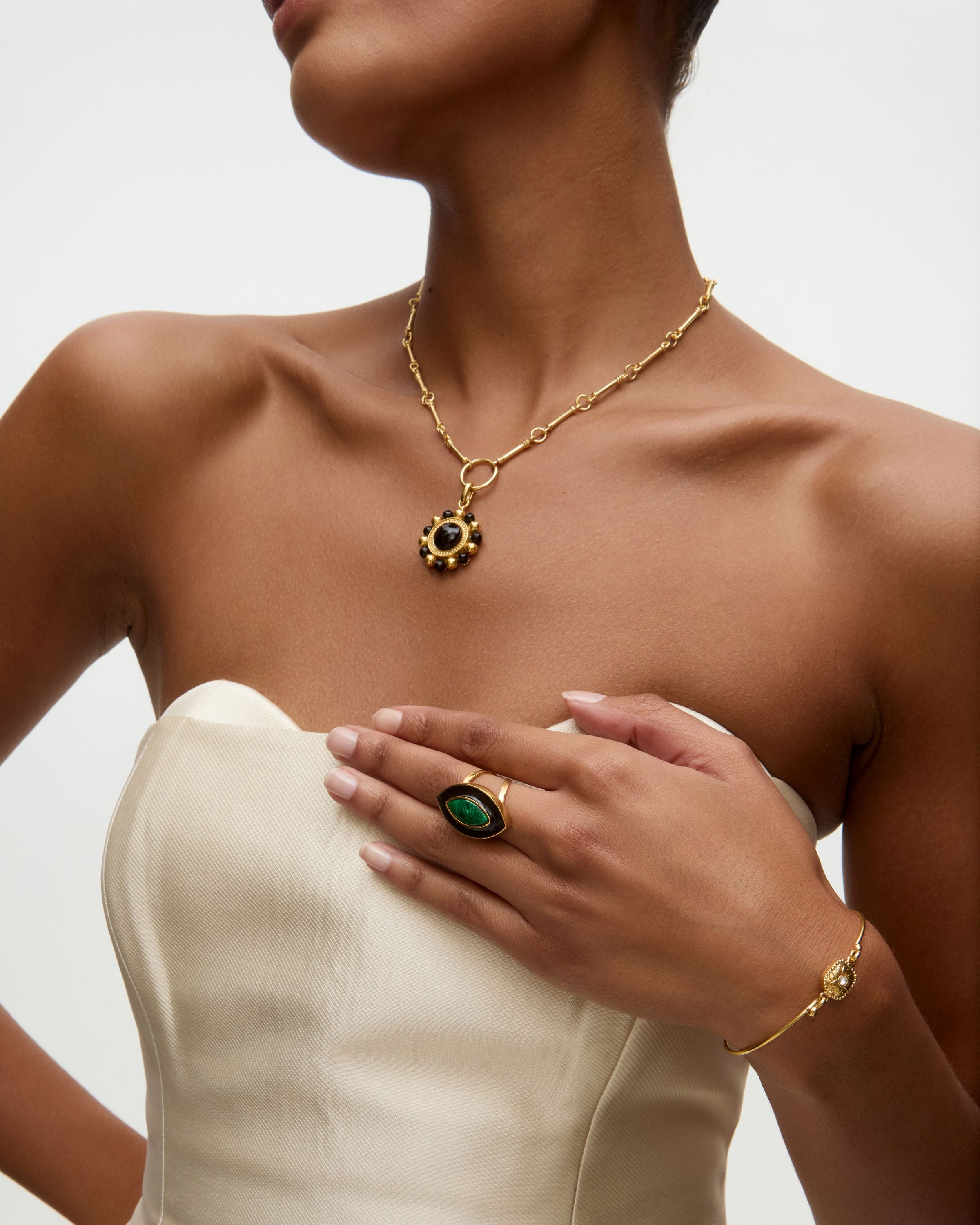 Woman wearing gold necklace and charm , green ring, and bracelet on a neutral background