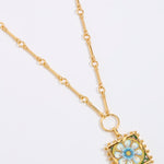 Gold square pendant with a floral tile design on a gold bar style chain on a white background 