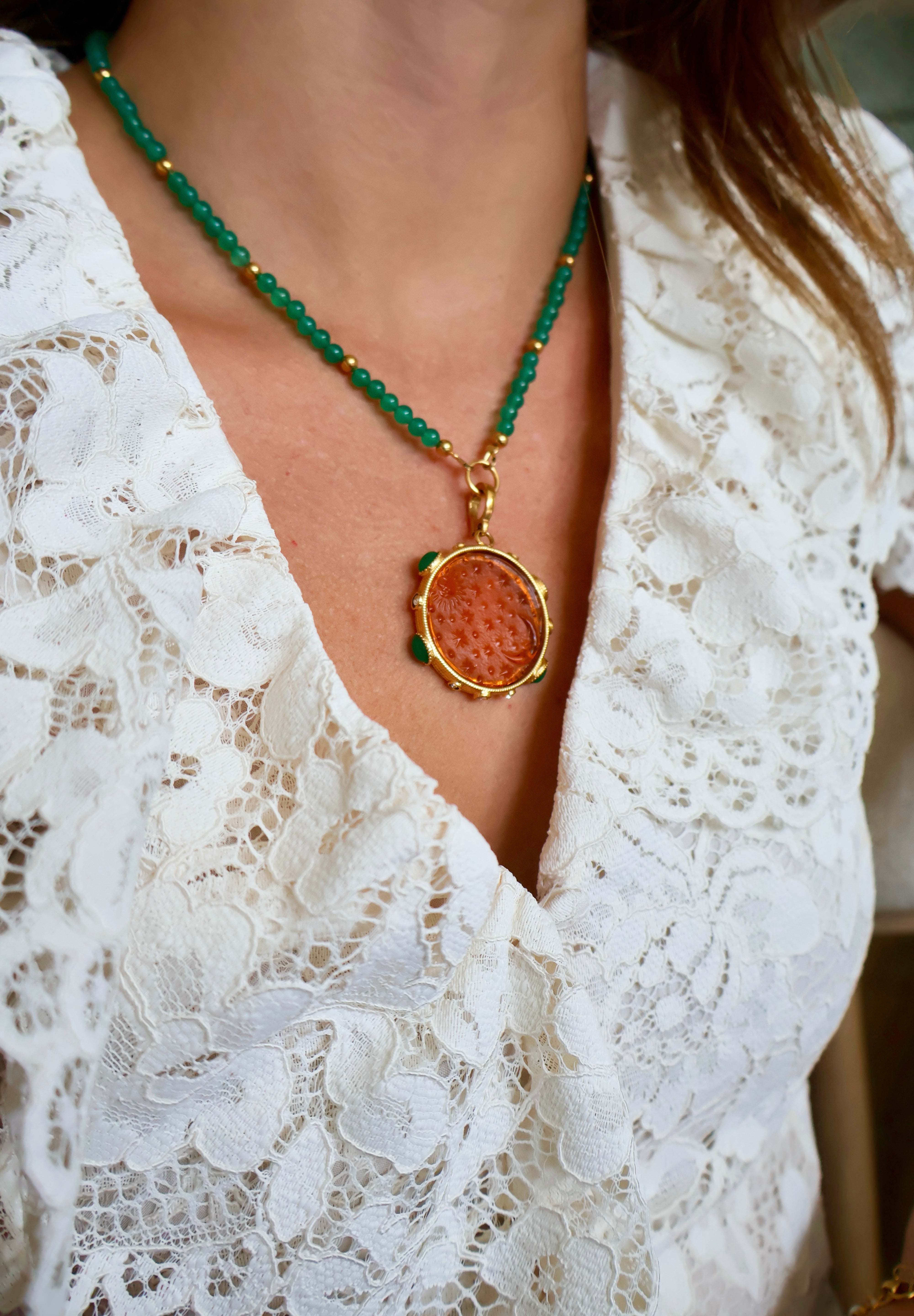 A close-up of a person wearing a  beaded necklace featuring round green gemstone beads separated by small gold spacer, and a round pendant featuring a textured orange-brown centre surrounded by a gold-tone border. The person is wearing a white lace top.