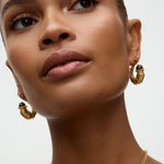 Close-up of a woman wearing gold hoop earrings and a necklace on a neutral background