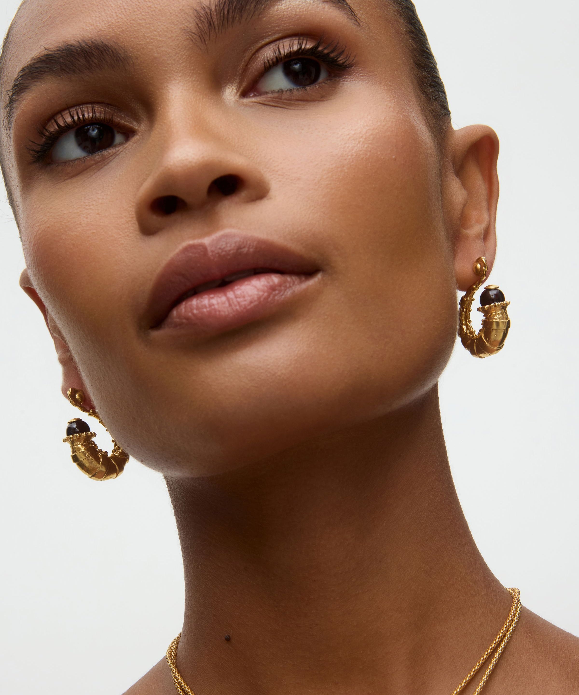 Close-up of a woman wearing gold hoop earrings and a necklace on a neutral background