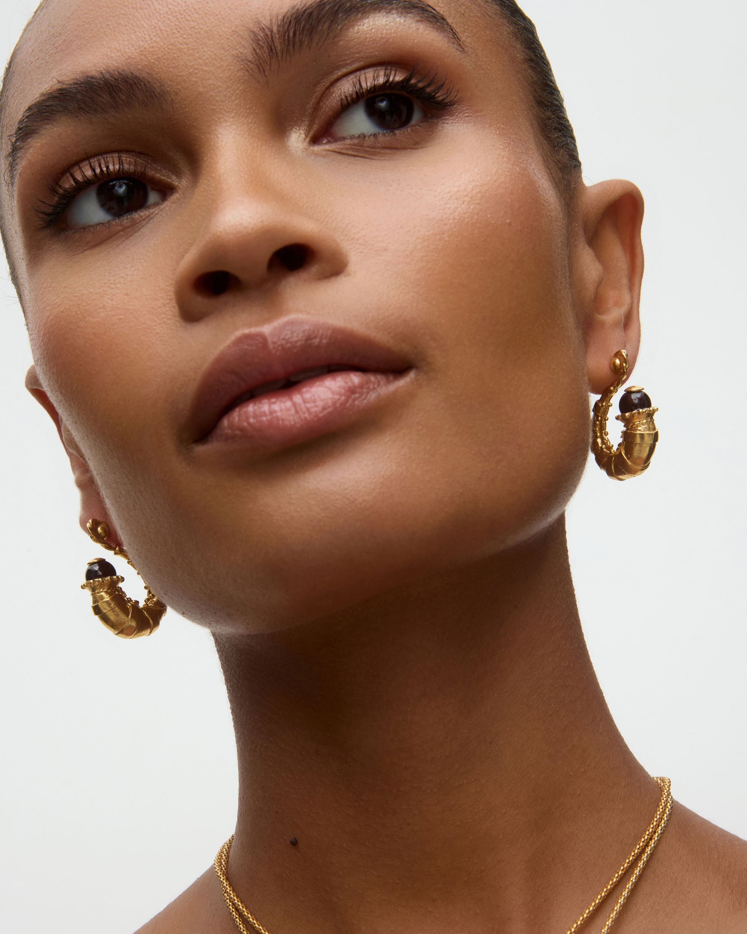 Close-up of a woman wearing gold hoop earrings and a necklace on a neutral background
